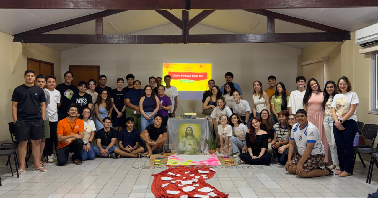 Grupo de jovens e adultos reunido durante encontro de preparação para a Crisma da Paróquia Cristo Rei, em torno de um espaço de oração com imagem de Jesus e materiais utilizados na atividade.