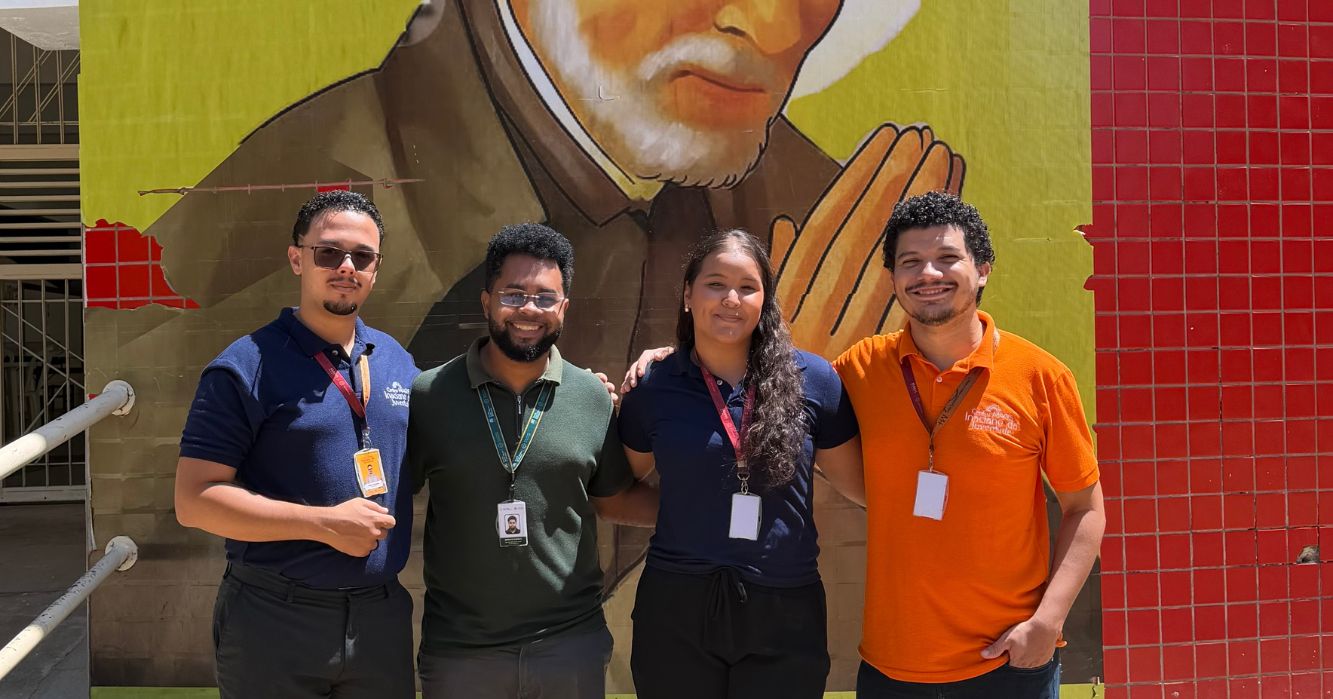 Quatro integrantes do Centro MAGIS Inaciano da Juventude posam juntos em frente a um mural religioso na Escola Santo Afonso Rodriguez, em Teresina/PI, durante atividade do Projeto Farol.