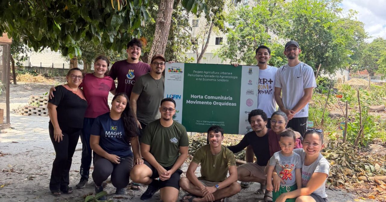 Grupo de participantes do ECOAR Ambiental posa ao lado da placa da Horta Comunitária Movimento Orquídeas, em Fortaleza. Jovens, colaboradores e parceiros aparecem reunidos em uma área verde, próxima aos canteiros da horta comunitária.