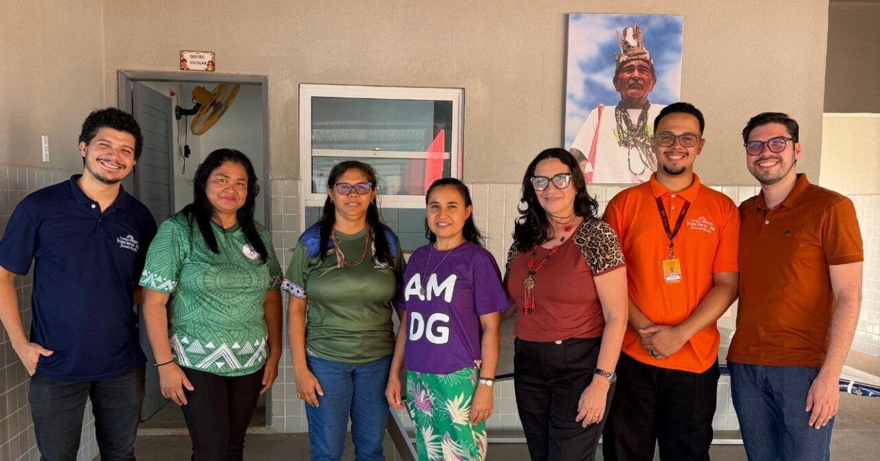 Sete pessoas posam lado a lado em um ambiente escolar, entre representantes do Centro MAGIS Inaciano da Juventude e membros da comunidade indígena Anacé. Ao fundo, há uma fotografia de uma liderança indígena e elementos do espaço da escola.