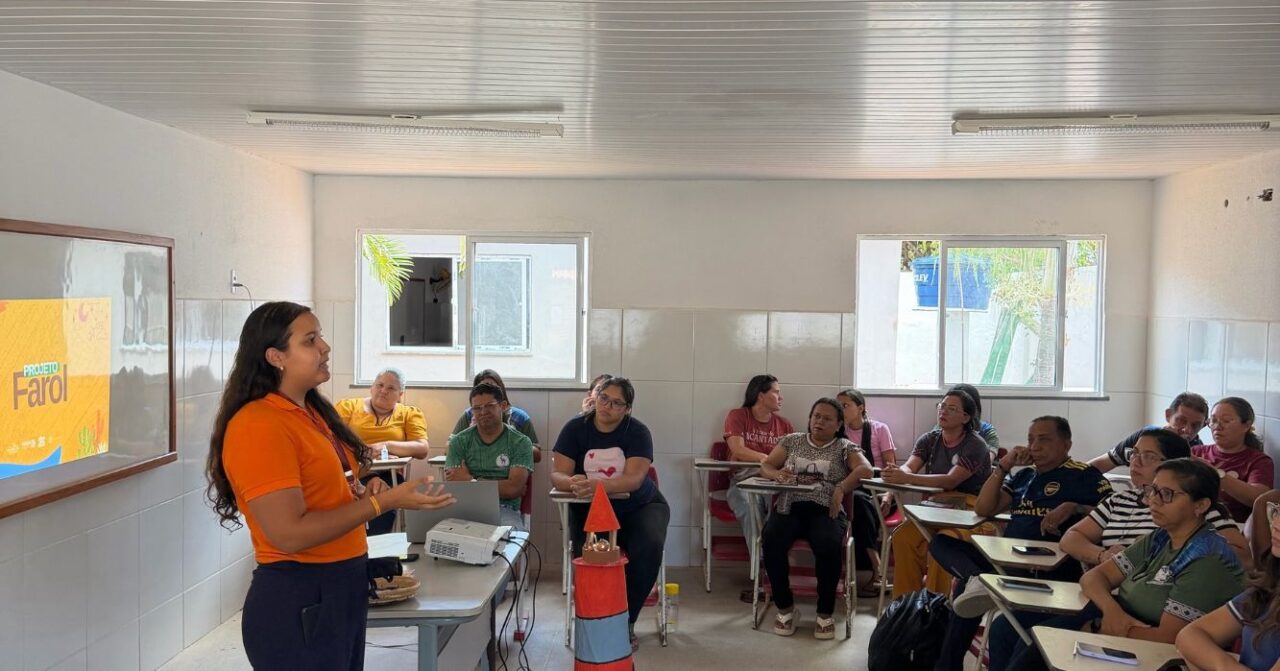 Mulher com camiseta laranja conduz uma apresentação do Projeto Farol em uma sala de aula. Participantes acompanham atentamente a atividade, sentados em carteiras, enquanto uma projeção com a identidade visual do projeto aparece na parede.